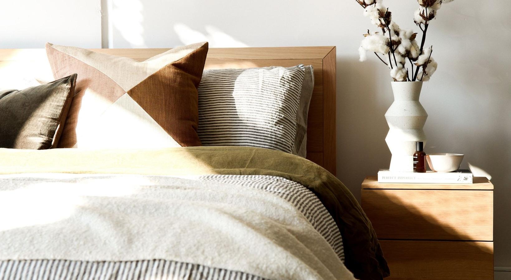 swath of sunlight shines across a well made bed and bedside table with a vase of cotton plants