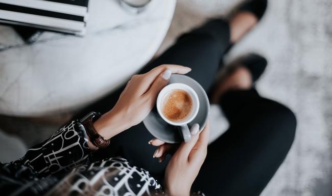 business woman sits next to a marble table with books and holds a saucer with a cup of espresso
