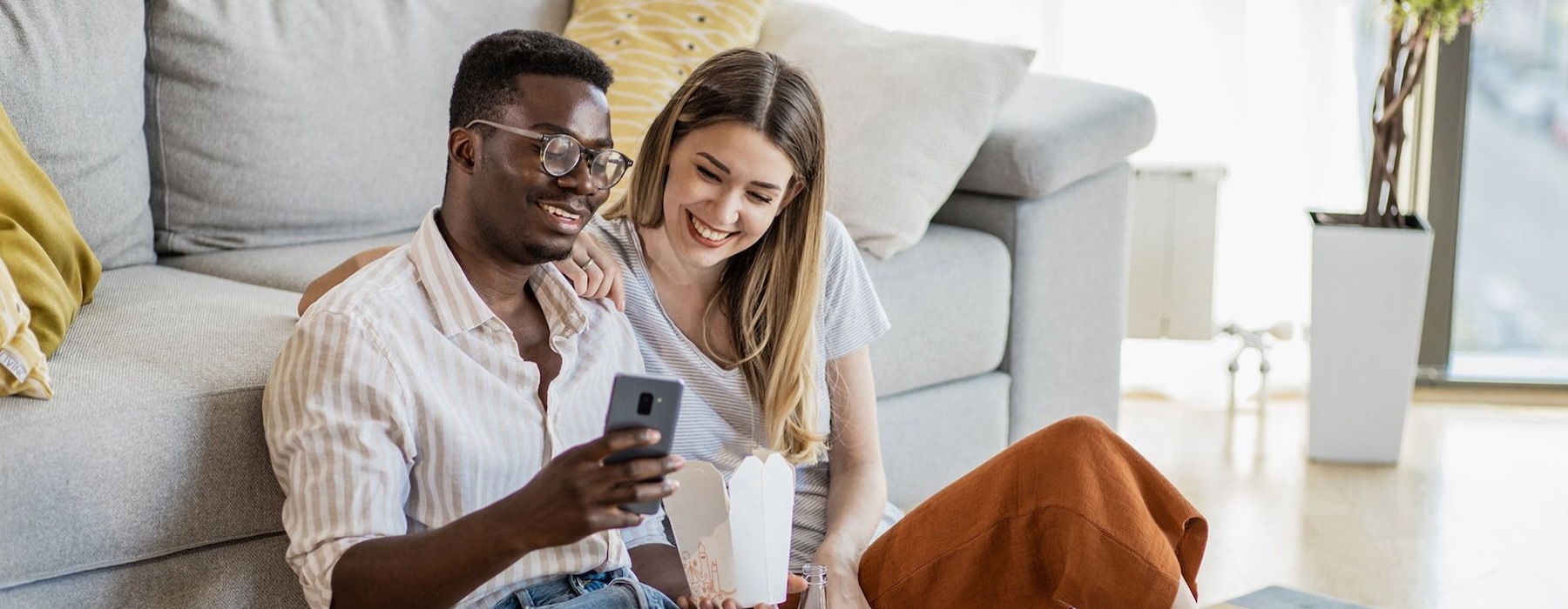 a man and woman with take out, sit against a couch on their living room floor and watch at their cell phone